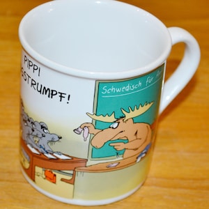 Puede incluir: Taza de cerámica blanca con una ilustración de dibujos animados. La taza presenta un alce y un ratón en un escritorio, con el texto "Pippi Strumpf!" y "Sueco para el arte". La taza tiene un asa curva.