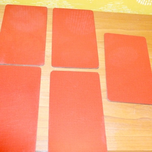 May include: Five rectangular, orange-red coasters are arranged on a light brown wooden surface. The coasters have a textured surface and rounded corners. The background features a yellow and white patterned wallpaper.