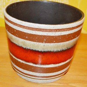 May include: A ceramic planter with a brown interior and a design of horizontal stripes in white, brown, and orange. The planter has a speckled texture and a slightly flared shape, suitable for indoor or outdoor use.