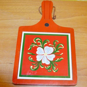 Pode incluir: Uma tábua de cortar de madeira com moldura e alça laranja. O centro apresenta um quadrado vermelho com um desenho floral branco, verde e azul. Uma alça de couro é anexada para pendurar.