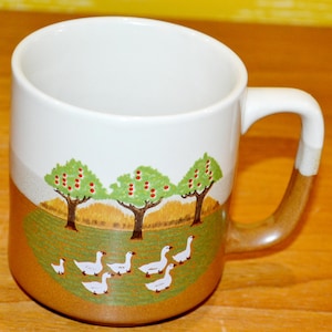 Puede incluir: Taza de cerámica blanca con asa y base marrón. La taza presenta una escena pastoral con árboles verdes con frutos rojos, un campo de hierba y gansos blancos. La taza tiene un diseño curvo.