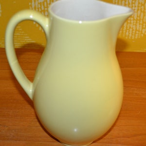 May include: A pale yellow ceramic pitcher with a rounded body and a handle. The pitcher is sitting on a wooden surface.