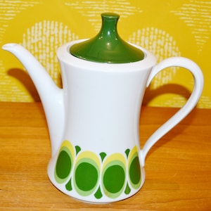 Könnte beinhalten: Weiße Keramik-Kaffeekanne mit grünem Deckel und gebogenem Griff. Die Kanne hat ein Retro-Design mit grünen und gelben ovalen Formen entlang der Basis. Der Ausguss ist lang und schlank.