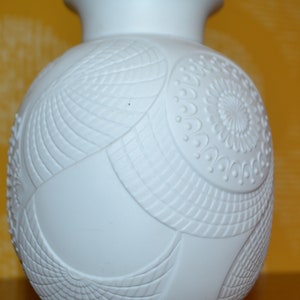 Puede incluir: Jarrón de cerámica blanca con un diseño en relieve intrincado. El jarrón tiene un cuerpo redondo y un cuello estrecho.