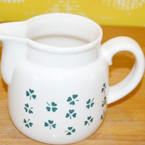 Puede incluir: Cremera de cerámica blanca con asa y boquilla, decorada con diseños de tréboles verdes. La cremera tiene un cuerpo redondeado y un asa curvada, perfecta para servir crema o leche.