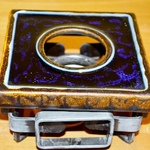 May include: Square ceramic trivet with a dark brown and blue glazed surface. The trivet has a circular opening in the center and a metal frame base. The edges are trimmed with a light blue and yellow border. A decorative kitchen item.