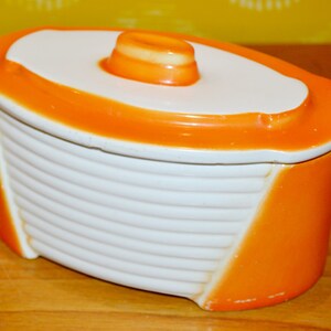 Könnte beinhalten: Orangefarbener und weißer Keramikbehälter mit Deckel. Der Behälter hat ein geripptes Design und eine abgerundete Form.