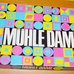 Könnte beinhalten: Vintage Brettspielbox mit einem farbenfrohen geometrischen Muster in Pink, Gelb, Blau und Schwarz. Der Spieletitel "MUHLE DAME" ist in großen weißen Buchstaben abgebildet. Die Box hat eine Retro-Ästhetik.