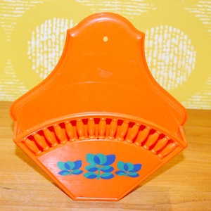 May include: Orange plastic wall-mounted coffee filter holder with a floral design. The holder has a scalloped edge and a hole for hanging.