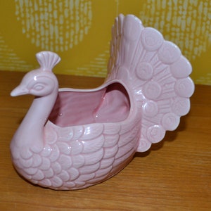 May include: A pink ceramic peacock planter with a detailed design of feathers. The planter has a hollowed-out space in the back for planting.