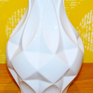 May include: A white ceramic vase with a geometric design. The vase has a wide base and a narrow neck.