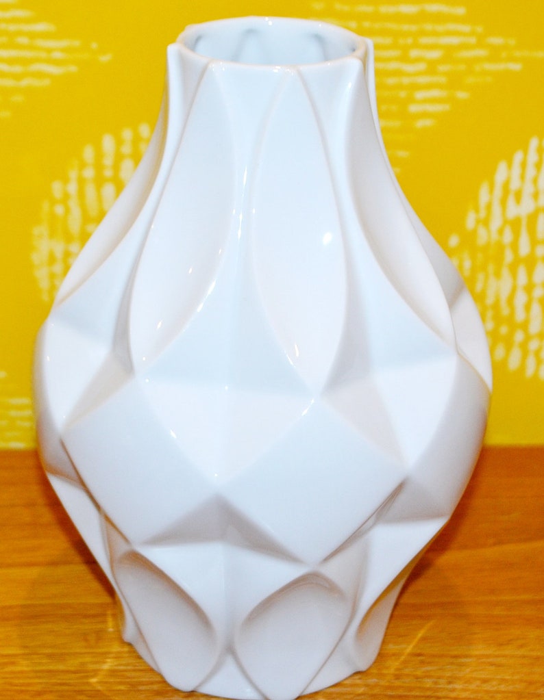 May include: A white ceramic vase with a geometric design. The vase has a wide base and a narrow neck.