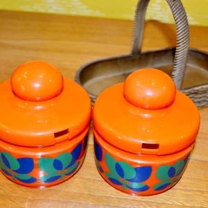 May include: Two orange ceramic condiment jars with blue and green leaf designs. The jars have lids with a small hole in the top. The jars are sitting on a wooden surface.