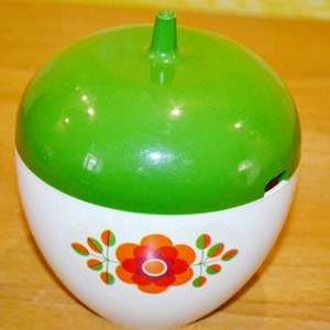 Op de afbeelding: Een witte keramische suikerpot met een groen deksel in de vorm van een appel. De pot heeft een bloemmotief met rode, oranje en witte bloemen.