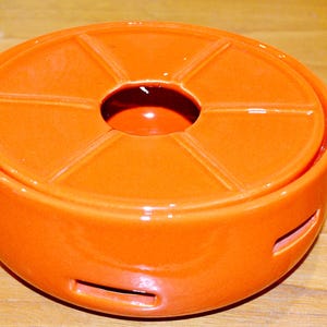 Könnte beinhalten: Ein orangefarbener Keramik-Fondue-Topf mit Deckel, der ein zentrales Loch und ein segmentiertes Design aufweist. Der Topf hat einen abgerundeten Boden mit mehreren rechteckigen Öffnungen. Die glänzende Oberfläche reflektiert das Licht.