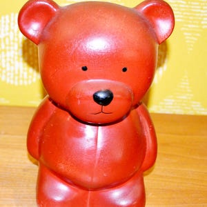 May include: A red ceramic teddy bear figurine with a glossy finish. The bear has small black eyes, a black nose, and a simple mouth. It stands upright with its arms at its sides and its legs together. The background is a yellow wall.