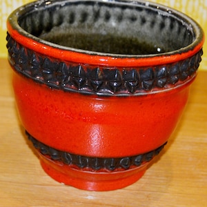 May include: A vibrant red ceramic planter with a textured black band around the top and bottom. The interior is dark, and the rim is outlined in black. The planter has a glossy finish and a unique, decorative design.