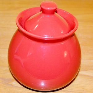 May include: A red ceramic sugar bowl with a lid. The bowl is round and has a smooth, glossy finish.
