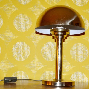 Könnte beinhalten: Eine goldene Tischlampe mit einem weißen Lampenschirm und einem runden Fuß. Die Lampe steht auf einer gelben, gemusterten Oberfläche.