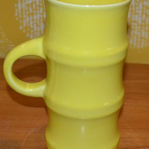 K&ouml;nnte beinhalten: Eine hohe gelbe Keramiktasse mit Bambus-Design. Die Tasse hat einen breiten Henkel und eine glatte, gl&auml;nzende Oberfl&auml;che.