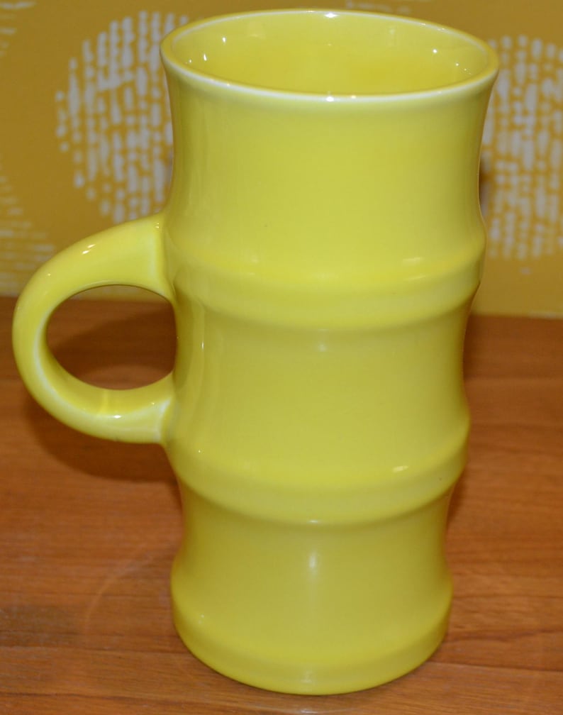 K&ouml;nnte beinhalten: Eine hohe gelbe Keramiktasse mit Bambus-Design. Die Tasse hat einen breiten Henkel und eine glatte, gl&auml;nzende Oberfl&auml;che.