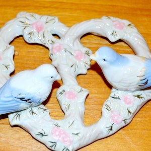Könnte beinhalten: Ein weißer Porzellan-Herz-Anhänger mit zwei blauen und weißen Vögeln, die darauf sitzen. Der Anhänger ist mit rosa Blumen und goldenen Akzenten verziert.