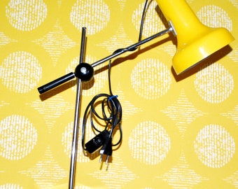 May include: A yellow desk lamp with a chrome arm and base. The lamp has a cylindrical head and a black power cord. The base is round and the lamp is set against a yellow patterned background.