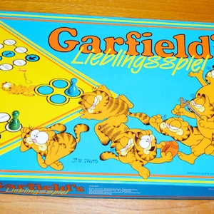 Könnte beinhalten: Vintage Garfield's Lieblingsspiel Brettspielbox. Die Box hat einen blauen Hintergrund mit orangefarbenem Text und Garfield-Illustrationen. Das Spielbrett hat ein gelbes und blaues Design mit Spielfiguren.