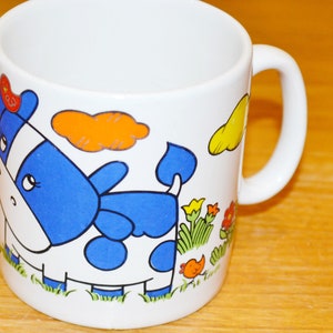 May include: White ceramic mug featuring a blue and white cow design. The cow has orange and yellow accents and is surrounded by flowers and clouds.