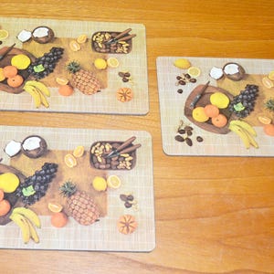 Pode incluir: Três tapetes de mesa com textura tecida apresentam uma natureza morta colorida de frutas, nozes e cocos. Os tapetes de mesa são bege com um padrão tecido castanho.