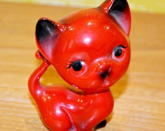 May include: A glossy red ceramic cat figurine with black accents on the ears and nose. The cat has large, expressive eyes with long eyelashes and a curled tail. The figurine is posed with its paw near its face.