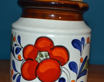 May include: A white ceramic jar with a brown lid and a floral design. The jar has a red, orange, and blue floral pattern with green leaves.