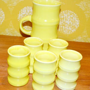 K&ouml;nnte beinhalten: Ein Set aus sechs gelben Keramikbechern und einem passenden Krug. Die Becher haben ein bambusartiges Design und sind perfekt zum Servieren von Getr&auml;nken.