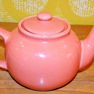 Könnte beinhalten: Eine rosa Keramik-Teekanne mit Deckel und Henkel. Die Teekanne hat einen abgerundeten Körper, einen gebogenen Ausguss und einen Schlaufengriff. Der Deckel hat einen kleinen Knopf oben. Die Teekanne steht auf einer Holzoberfläche.