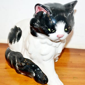 Könnte beinhalten: Eine Keramik-Katzenfigur in sitzender Position, mit einem schwarz-weißen Tuxedo-Muster. Die Katze hat grüne Augen, eine rosa Nase und schwarze Ohren. Die Figur steht auf einer Holzoberfläche.