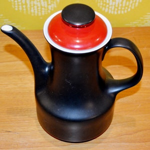 May include: Black ceramic coffee pot with a red and black lid. The pot has a rounded body and a spout on the side. The lid has a white rim.