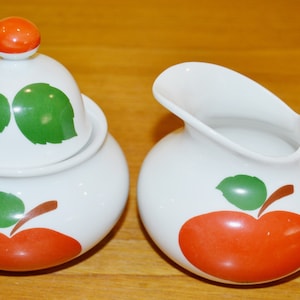 Peut inclure: Un ensemble de pot à crème et de sucrier en céramique blanche avec un motif de pomme rouge et de feuille verte.
