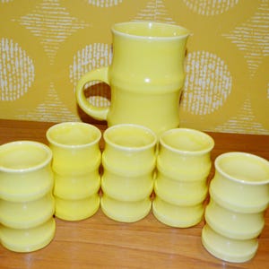 K&ouml;nnte beinhalten: Ein Set aus sechs gelben Keramikbechern und einem passenden Krug. Die Becher haben ein geriffeltes Design und der Krug eine glatte Oberfl&auml;che.