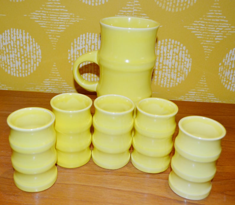 K&ouml;nnte beinhalten: Ein Set aus sechs gelben Keramikbechern und einem passenden Krug. Die Becher haben ein geriffeltes Design und der Krug eine glatte Oberfl&auml;che.