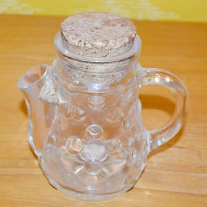 May include: A clear glass pitcher with a cork stopper. The pitcher has a textured surface with small raised circles. The pitcher has a handle and a spout.