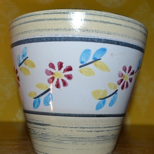 May include: A white ceramic planter with a yellow, blue, and red floral design. The planter has a textured, cream-colored rim and base.