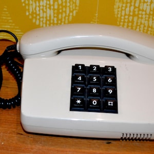 Könnte beinhalten: Cremefarbenes Vintage-Telefon mit schwarzer Tastatur mit Zahlen und Symbolen. Der Hörer liegt auf der Basis und ist über ein Spiralkabel verbunden. Das Telefon steht auf einer Holzoberfläche mit gelbem Hintergrundmuster.