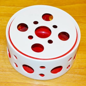Könnte beinhalten: Weißer Keramik-Tee-Wärmer mit rotem Rand und roten Kreisen, die an den Seiten ausgeschnitten sind. Die Oberseite des Wärmers hat einen roten Kreis in der Mitte und kleinere rote Kreise darum herum.