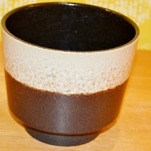 May include: A ceramic planter with a three-tone design. The interior is black, the middle section is off-white, and the base is dark brown. The planter has a slightly angled shape and a small, circular base.