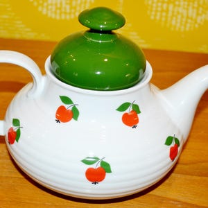 Peut inclure: Une théière blanche avec un couvercle vert et des motifs de pommes rouges. La théière a un corps rond et un bec verseur sur le côté.