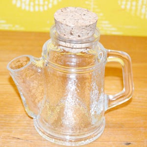 May include: A clear glass creamer with a textured surface, featuring a spout and handle. It is topped with a cork stopper. The creamer is designed to hold liquids and is suitable for serving.