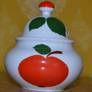 Peut inclure: Sucrier en céramique blanche avec un motif de pomme rouge et verte. Le couvercle a un bouton rouge et deux feuilles vertes.