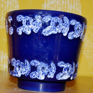 May include: A blue ceramic planter with a white, textured design. The planter has a wide, round base and a slightly flared rim.