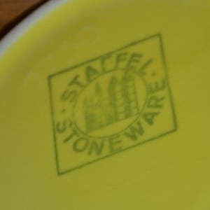 K&ouml;nnte beinhalten: Ein gr&uuml;ner Stempel auf gelbem Hintergrund mit dem Text "Stafford Stoneware" und einem stilisierten Bild eines Geb&auml;udes.