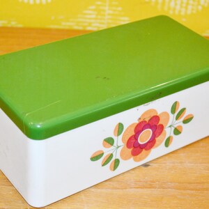 Puede incluir: Una caja rectangular blanca con tapa verde brillante. La caja presenta un diseño floral en tonos naranja, rojo y verde. La caja es probablemente de plástico y adecuada para el almacenamiento.